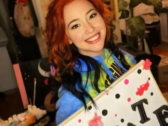 Smiling red-haired Rachel in a colorful tie-dye hoodie holding a decorated sign indoors with plants and string lights behind her.