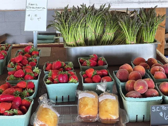 Farm stand table displaying Harris strawberries, fresh asparagus, South Carolina peaches, and packaged baked goods with handwritten price signs.
