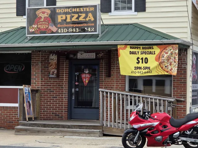 Storefront of Dorchester Pizza in Secretary, Maryland, with a green awning, a crab holding a pizza slice on the sign, an open sign, and a banner advertising happy hour specials.