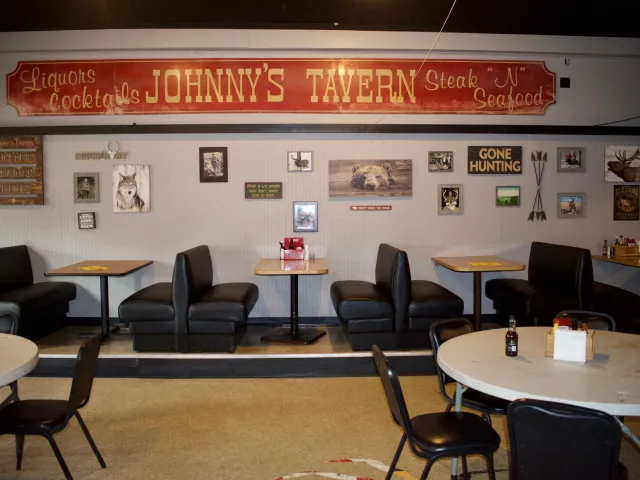 Interior of Johnny's Tavern in East New Market, Maryland, with a vintage red Johnny's Tavern sign reading Liquors Cocktails and Steak N Seafood above a row of booths, hunting-themed wall art, and round tables.